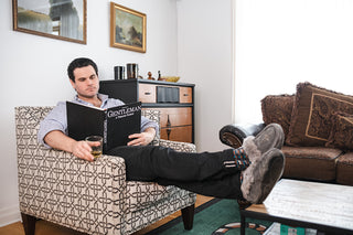 A man is enjoying a book and a drink on the sofa in The Tiger's Den, the main living room of Cellar House NOTL.